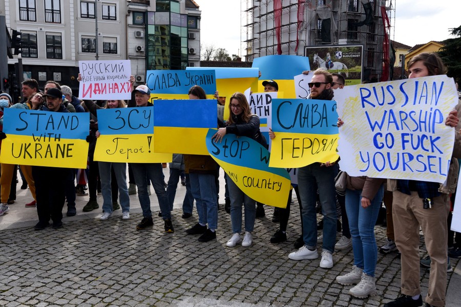 A small crowd of people stands in a plaza holding protest signs, including one that reads "Russian warship go fuck yourself."