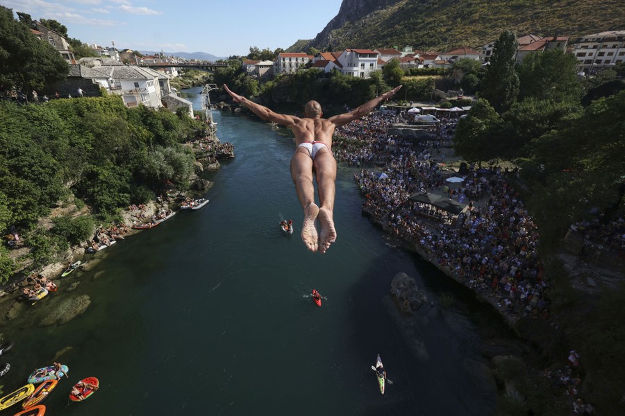 A diver leaps from a bridge high above a river, as a crowd of observers watch below.