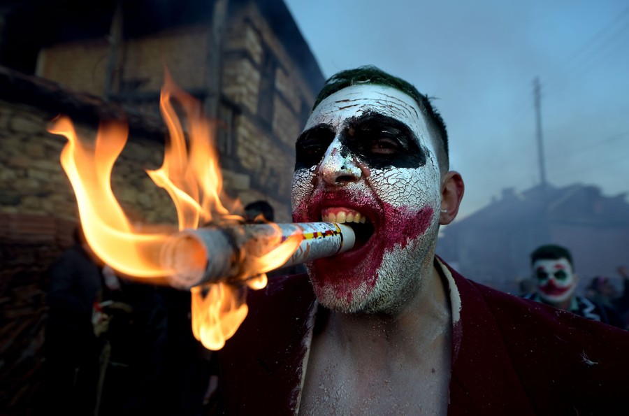 A man wearing cracked clown makeup holds a flaming tube in his teeth, as others parade beside him.