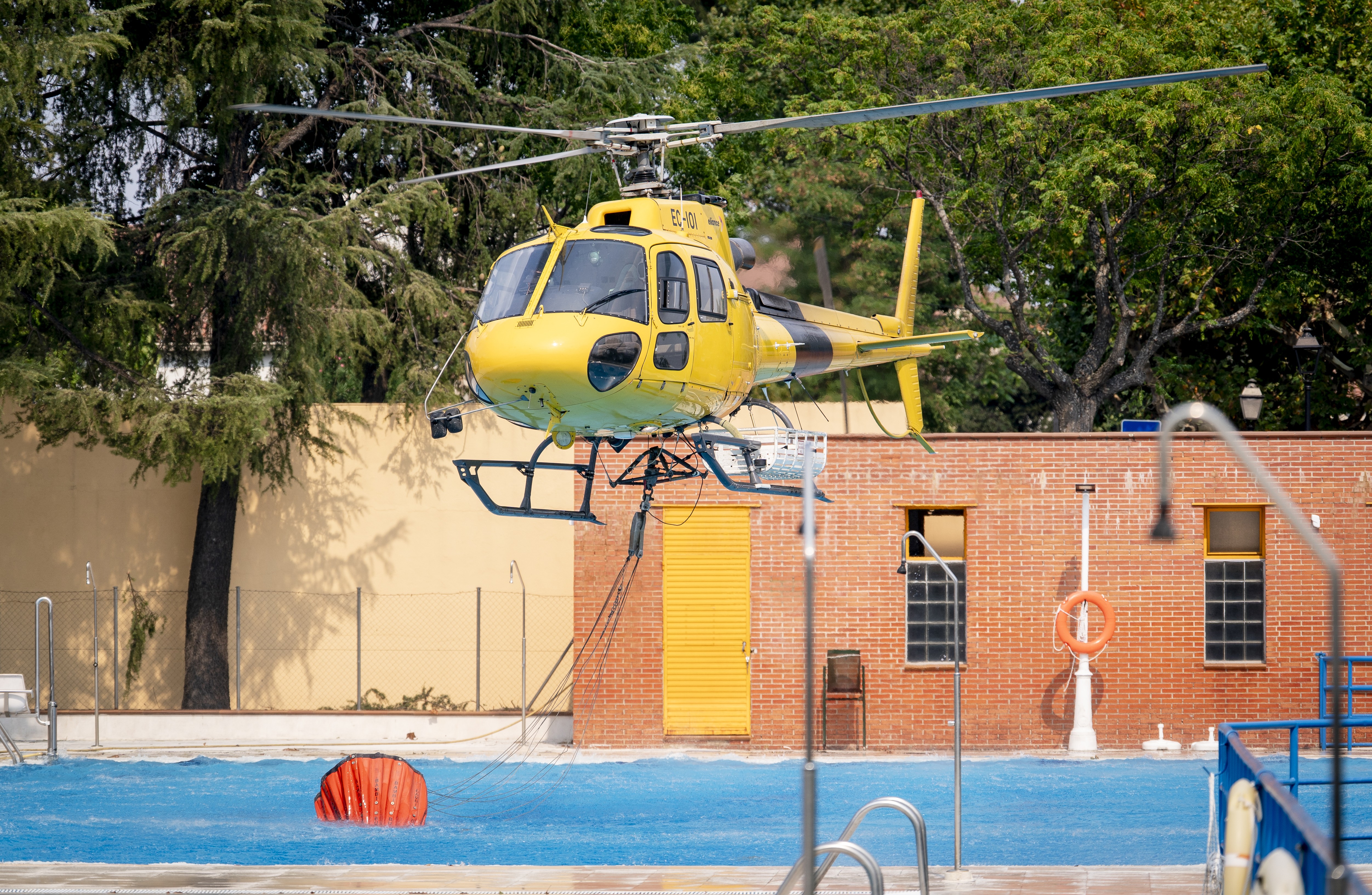 A firefighting helicopter hovers just above a swimming pool as it uses a bucket to scoop up water.