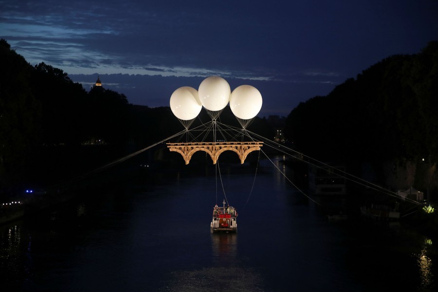 Three large balloons hold up a sculpture of a bridge above a river at night.