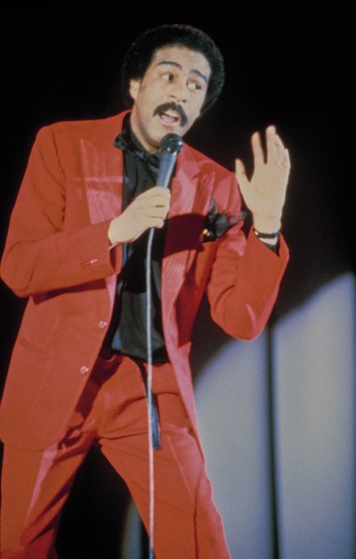 photo of Richard Pryor wearing red suit and black shirt performing on stage with mic