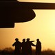 Photograph of black silhouettes of military personnel beneath the wing of a fighter jet as the sun shines brightly behind them