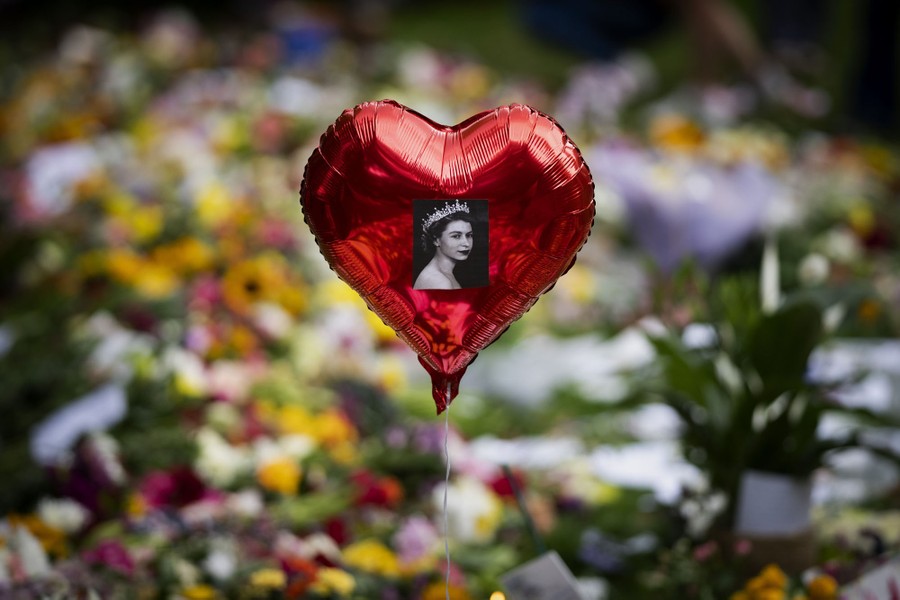 A heart-shaped balloon with a picture of Queen Elizabeth II floats near piles of flowers.