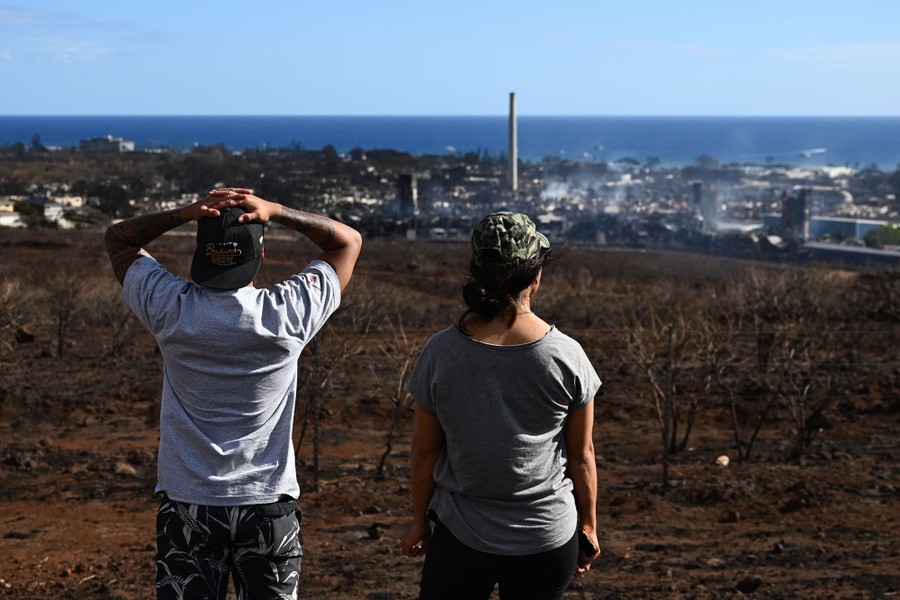 Two people look toward the distant, still-smoking ruins of a town.