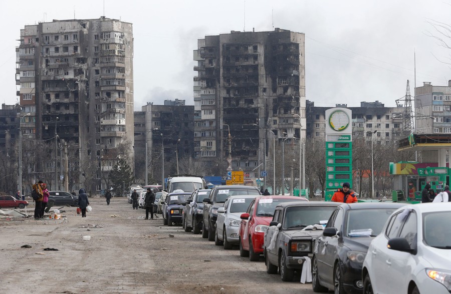 Uma longa fila de carros fica em uma rua, com vários prédios de apartamentos queimados atrás deles.