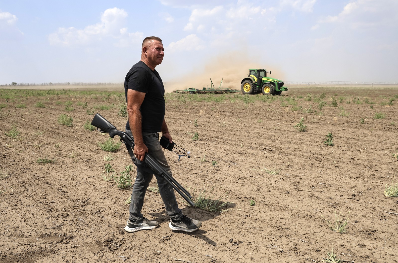 A person walks in a farm field, carrying a long gun and a hand-held electronic device, watching for drones, as a farm worker drives a tractor in the background.