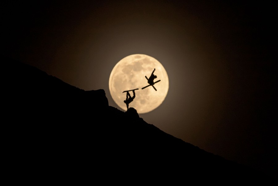 A skier and snowboarder perform tricks together, silhouetted by the full moon.