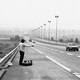 A black and white photograph of a hitchhiker with two bags facing away from the camera standing at the side of the road while a small car approaches on the far lane