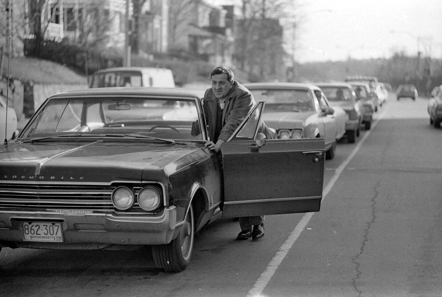 A man stands beside his car, pushing it with the door open.