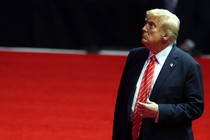 Donald Trump touches his tie and looks up while standing on a red carpet