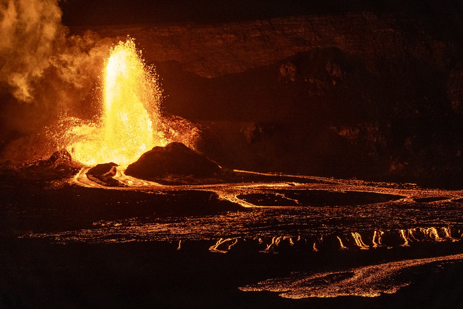 A fountain of lava blasts from a crater, then flows out in several small streams of glowing molten rock.