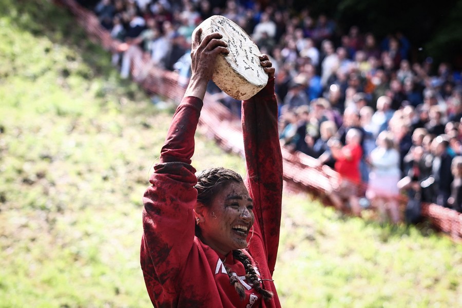 A woman, covered in mud splatters, holds a round wheel of cheese over her head, celebrating her win.