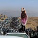 A woman holds her arms up, gesturing, while standing on a car among a large crowd of people marching past cars on a major road.