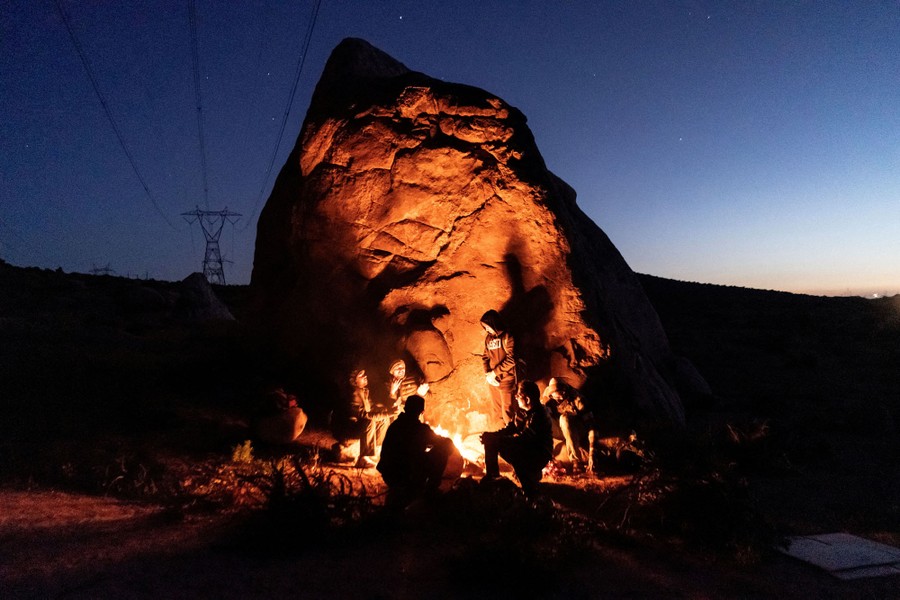 A half-dozen people sit around a campfire beside a tall rock in the desert at night.