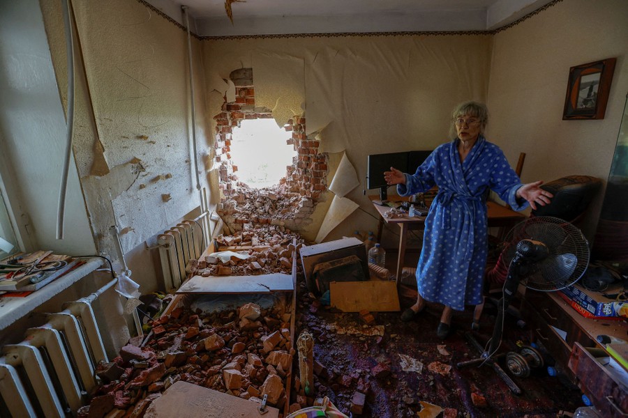 A person stands in a wrecked apartment, gesturing near a hole blasted in the outer brick wall.