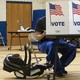 A child in a carrier sits next to their father as he votes. The father's face is covered by a partition that reads "VOTE" and is decorated with an American flag.