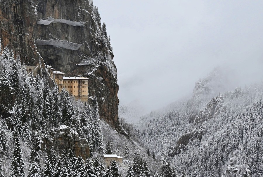 An ancient monastery built along a sheer and high cliff, above a snow-covered forested valley