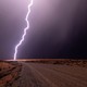 A single bolt of lightning in a dark sky illuminates arid ground beside a gravel road.