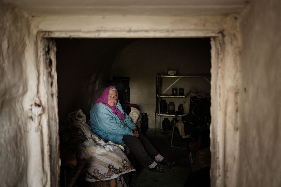 An older woman sits on a bed in a dark cellar, seen through a doorway.