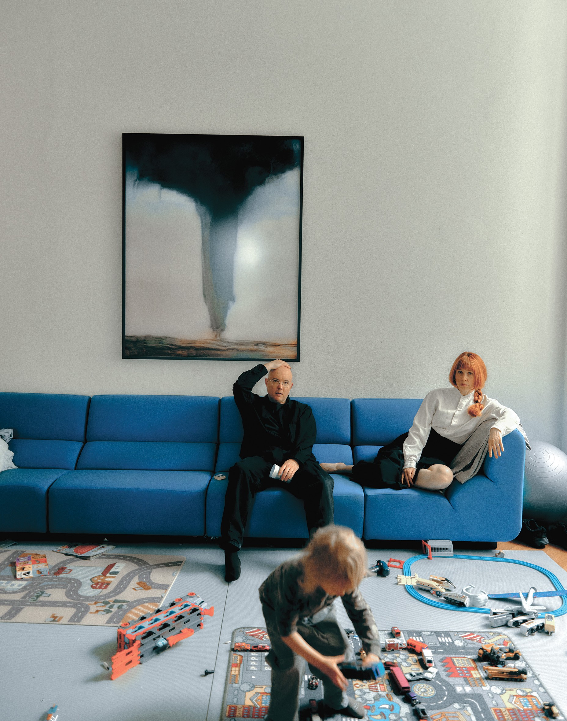 photo of bald man and red-haired woman sitting on long modern blue sofa with large abstract artwork on wall behind and young child playing on floor with toy vehicles in foreground