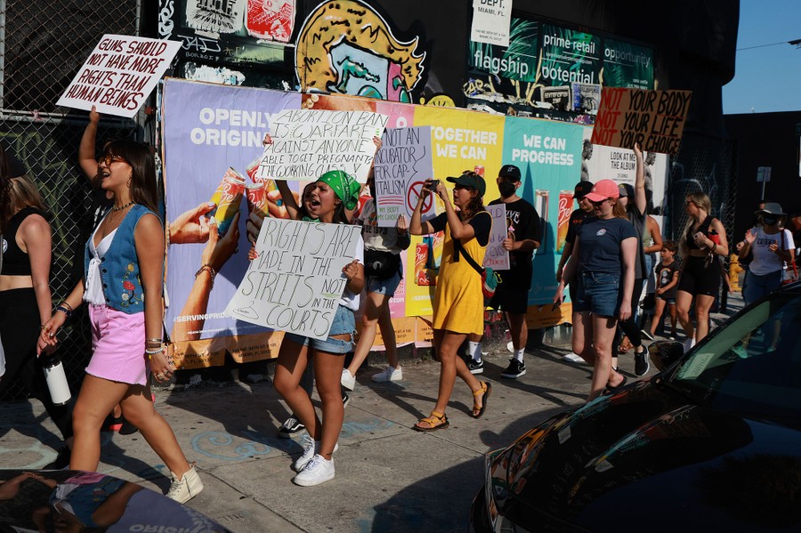Protesters carry signs while marching along a city sidewalk.