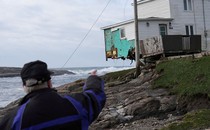 A person points toward a storm-damaged house hanging above a rocky shoreline.