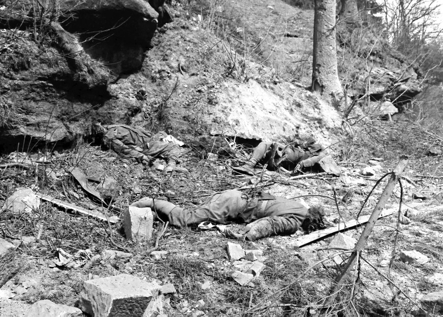 Three German soldiers lie dead among debris beside a road.