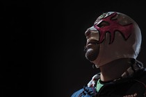 A color photograph of a man wearing a skin-tight white-and-pink luchador mask, set against a black background.