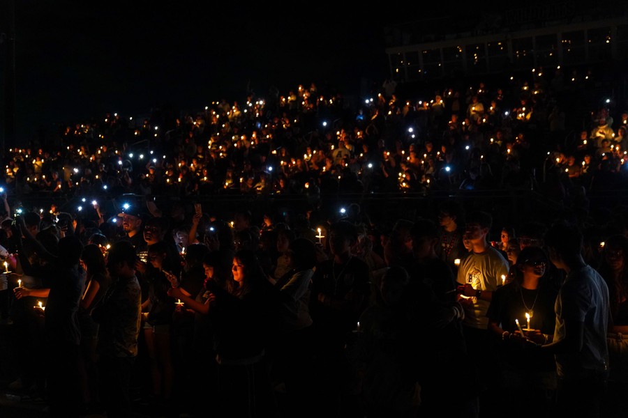 A crowd gathers near a high-school sports field, holding candles during a memorial.