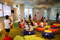 A group of elementary-school aged students sits on colorful chairs while looking at a powerpoint presentation. One boy holds a microphone and appears to be presenting.