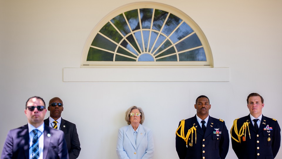 A photograph of Susie Wiles in a light suit and sunglasses, surrounded by military personnel and body guards