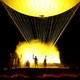 Five torchbearers gaze at a large gondola-like cauldron, below a large balloon, silhouetted by illuminated smoke.