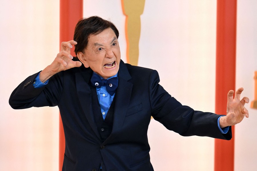 An actor, wearing a bowtie with googly eyes, strikes a fighting pose for cameras on a red carpet.
