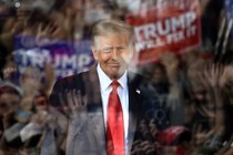 Donald Trump smiles behind a window of bulletproof glass that reflects his cheering supporters and signs