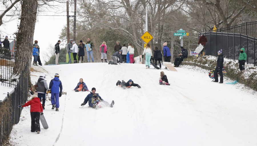 Several people sled down a snow-covered street, as a couple of dozen bystanders watch.
