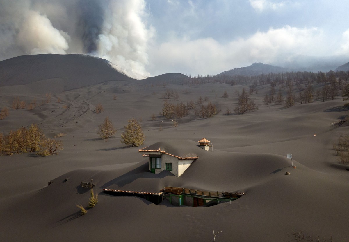 A house is nearly completely covered by ash, seen on a large ash-covered hillside beneath an erupting volcano.