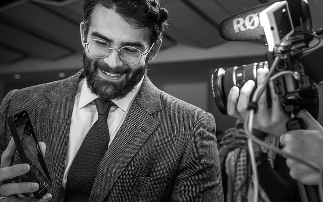 Hasan Piker is photographed smiling and holding his phone screen to the camera.