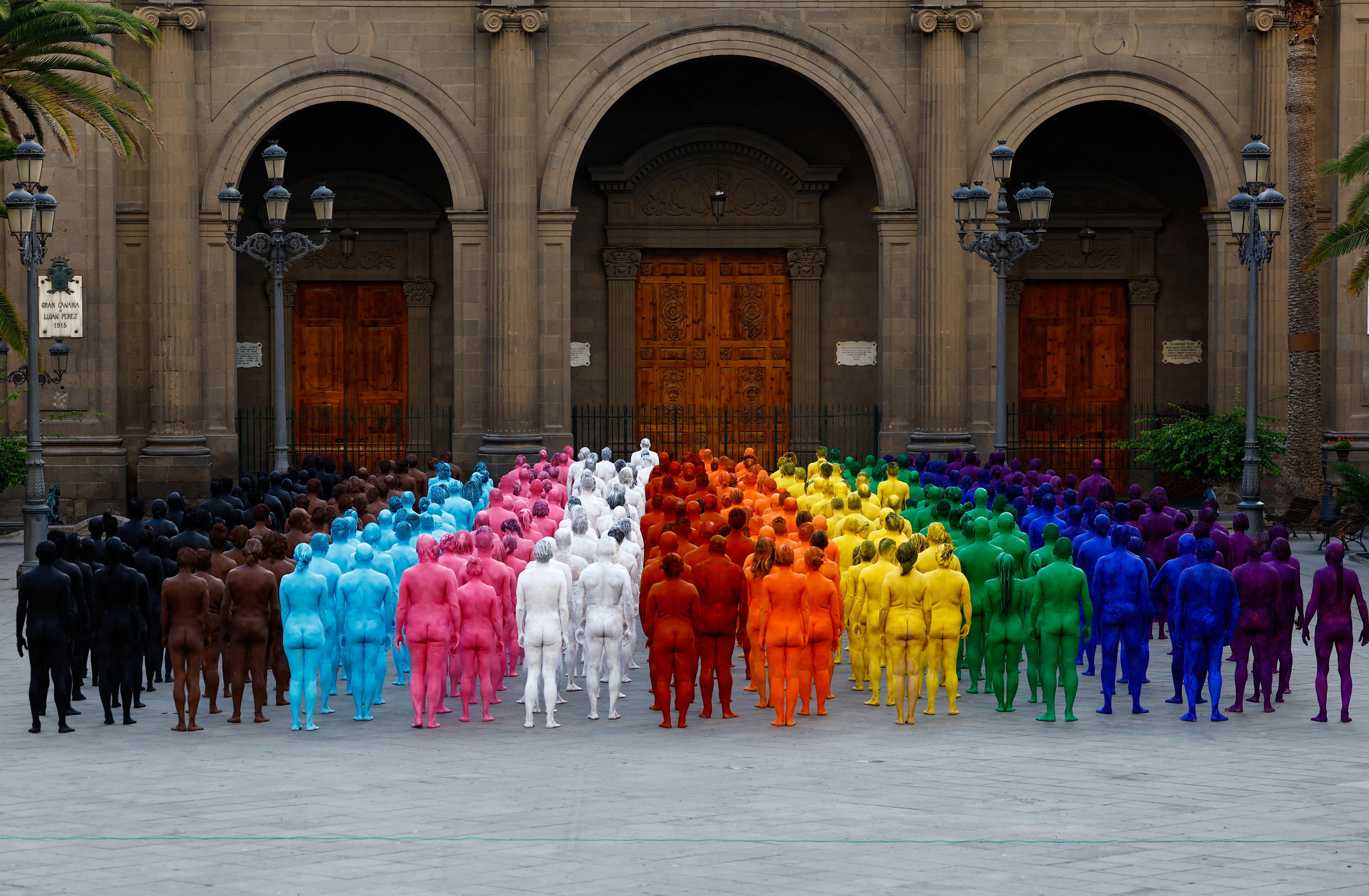 Dozens of people, nude and painted in a variety of colors, stand in a public square, forming rows of color, like a rainbow.