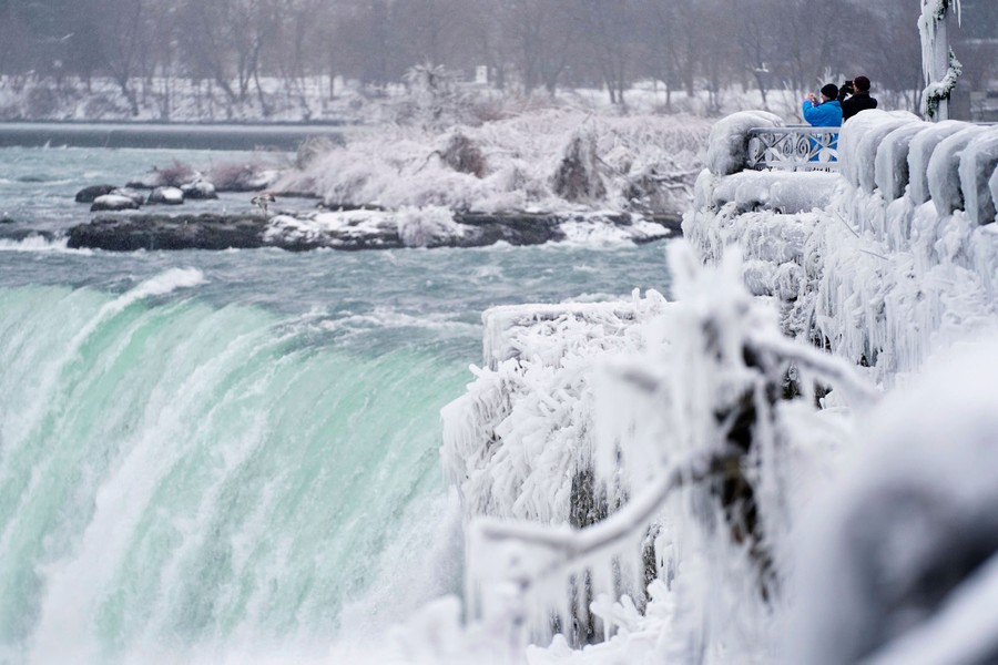 Two people stand along an ice-covered walkway beside rushing water going over Niagara Falls.