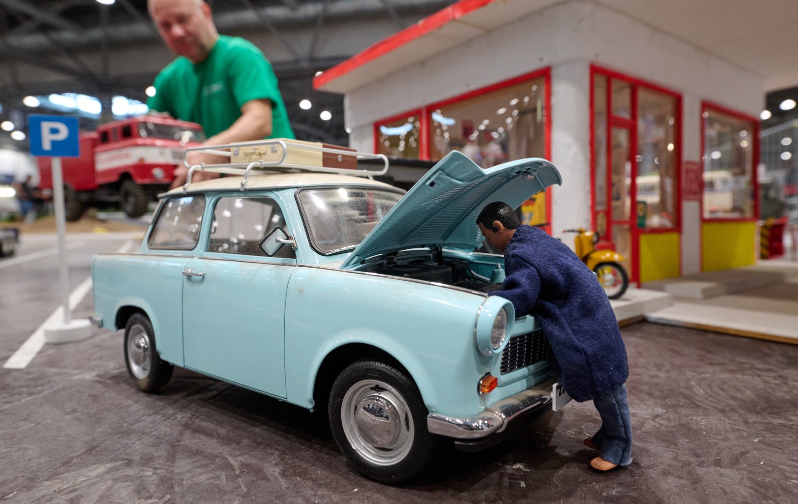 A close view of a miniature scale model of a person looking under the hood of an older car.