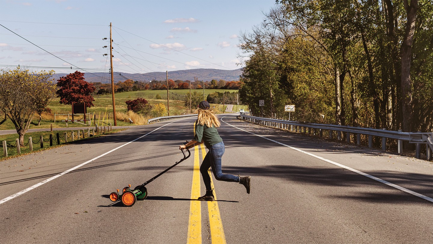 photo of woman with long hair pushing hand mower across rural two-lane road with mountains in background