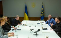 photo of large white meeting table with two women and a man on left and two men on right, one speaking, with Ukrainian flags in background