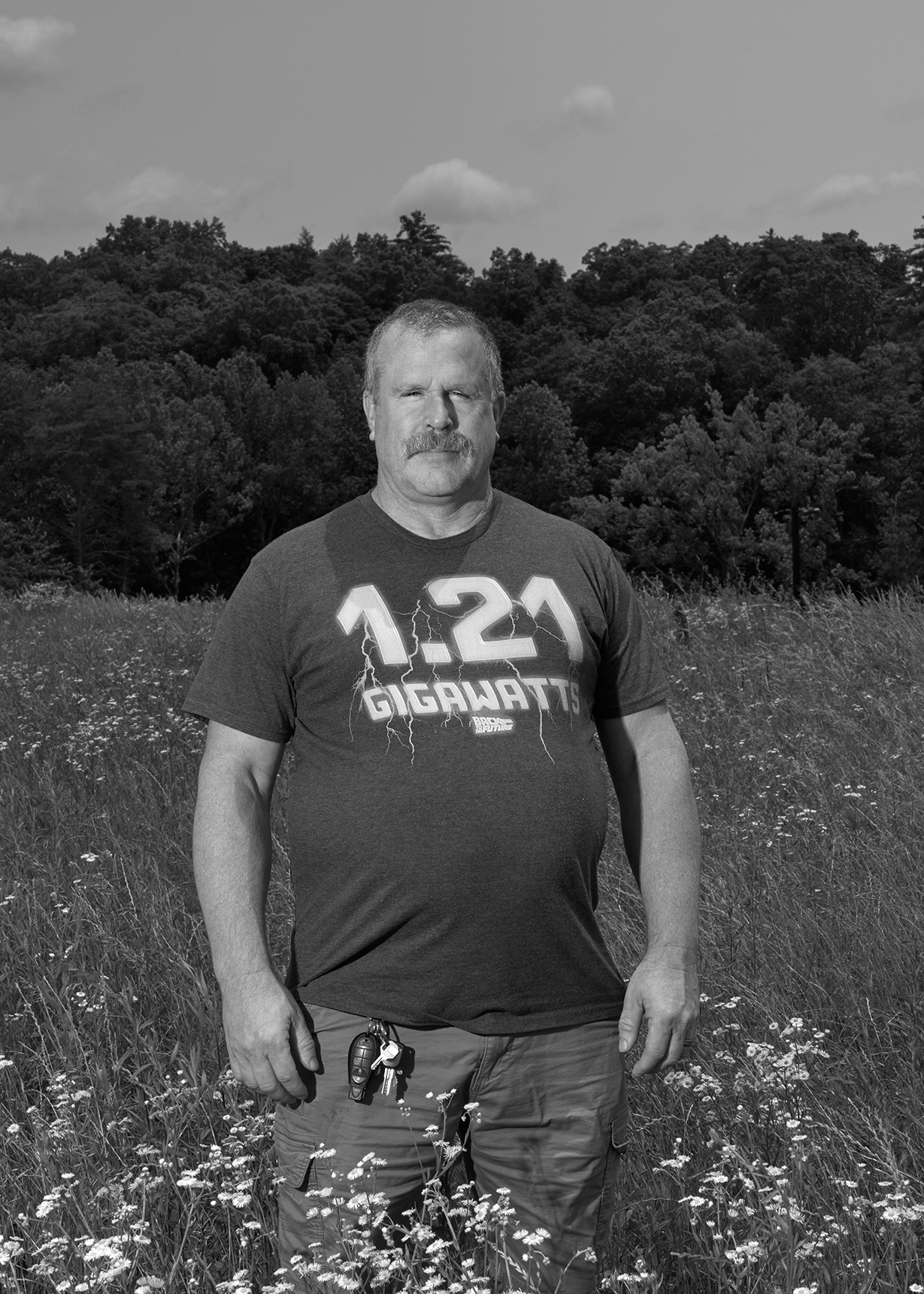 black-and-white photo of man wearing t-shirt with '1.25 Gigawatts' on it and pants standing in field of yellow flowers