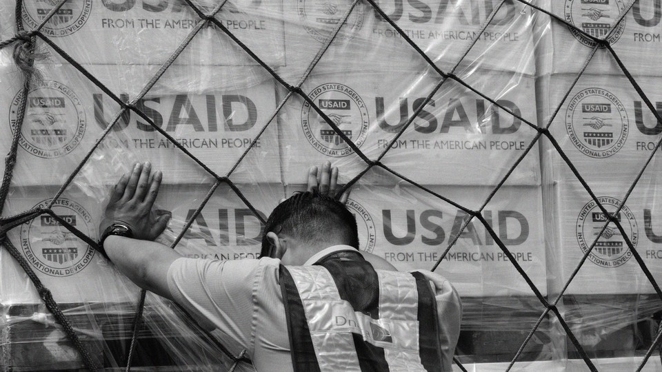 Black-and-white photograph of a person leaning against a stack of boxes labeled "USAID"