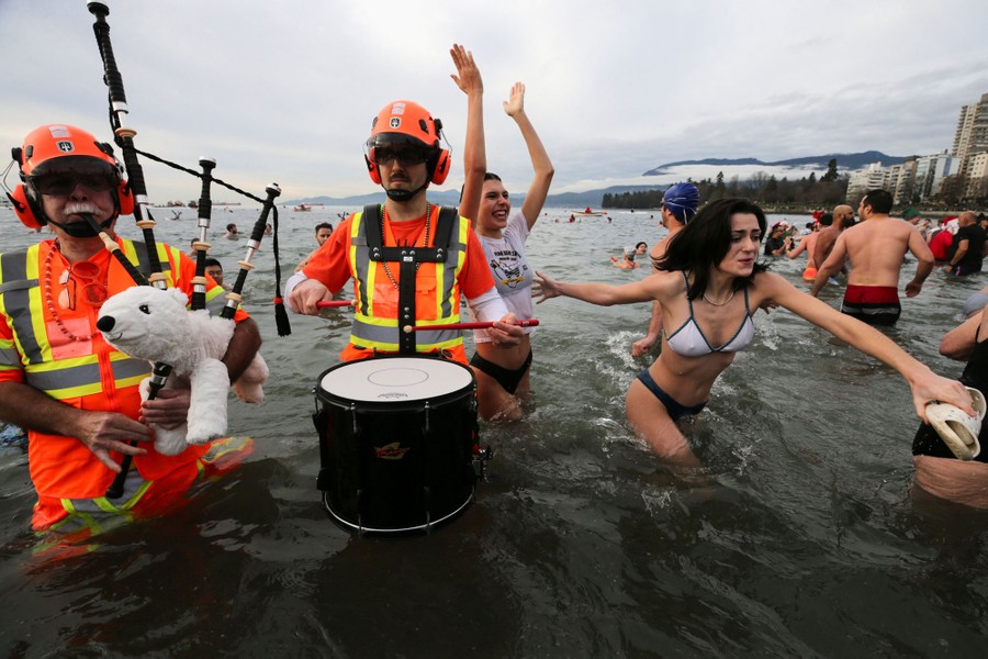 Two costumed people play a drum and a polar-bear-shaped bagpipe as swimmers play in waist-deep water along a beach.