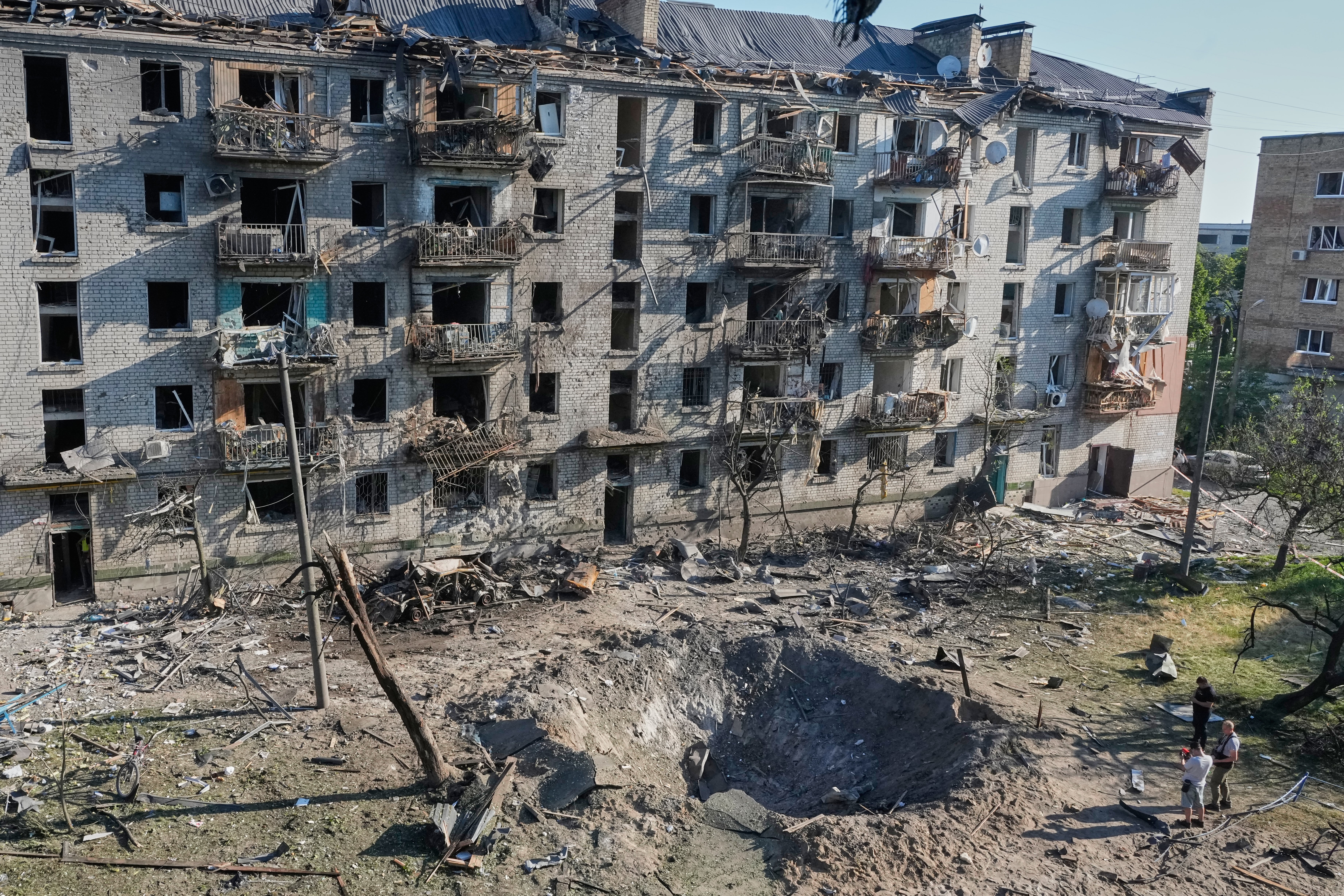 People look at a crater beside a heavily damaged apartment building.