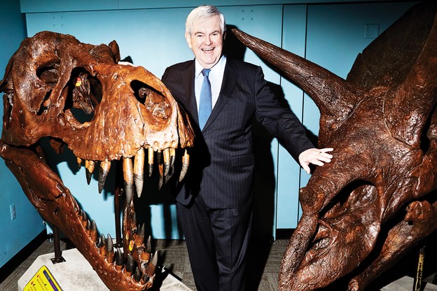 Newt Gingrich stands, grinning, between exhibits of dinosaur skeletons.