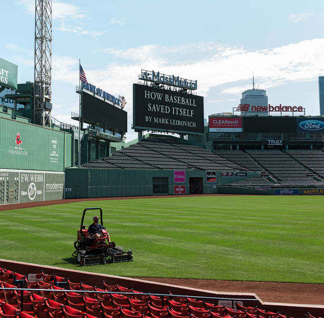 Man mowing the lawn at Fenway Park with the words "How Baseball Saved Itself by Mark Leibovich" behind him on the Jumbotron 