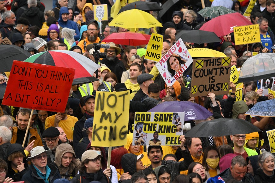 A crowd of protesters holds umbrellas and signs, reading 'Not my King' and 'Don't you think this is all a bit silly?'